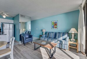 Un salon comprenant un canapé, un fauteuil et une table basse, une cuisine avec un réfrigérateur, et un coin repas avec des chaises et une table.