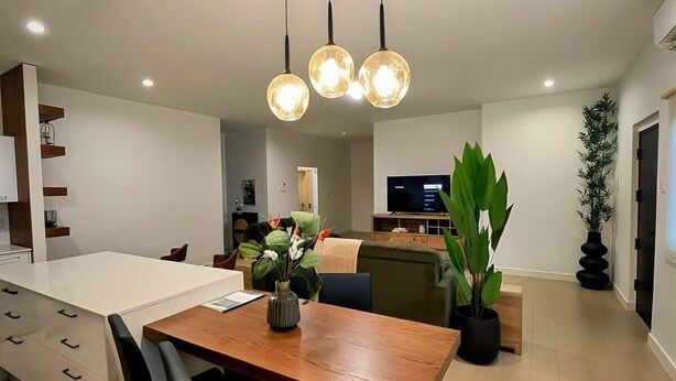 Een moderne keuken met een centraal kookeiland, hanglampen, een eethoek met stoelen, en een woonkamer met een bank en televisie.