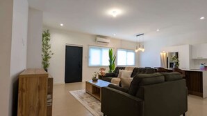 Amplia sala de estar con sofá, mesa de centro y una planta. Zona de cocina con armarios y un refrigerador.