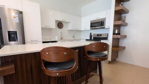 Una cocina moderna con armarios blancos, horno empotrado, microondas y dispensador de agua. Hay dos taburetes de bar de madera con asientos acolchados.