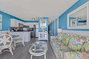 Ein Wohnzimmer mit einer Couch, Sesseln, einem Glastisch und einer Küchenzeile mit Geräten.