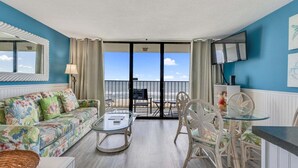 Quarto com sofá, mesa de vidro, cadeira e vista para a praia.