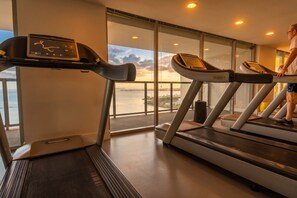 Une salle de sport équipée de tapis de course et d’un écran numérique, de grandes fenêtres offrant une vue sur la mer, et une personne utilisant un tapis de course.