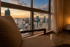 Ein Zimmer mit großem Fenster und Blick auf die Stadt, ein gepolsterter Sessel sowie ein kleiner Nachttisch.