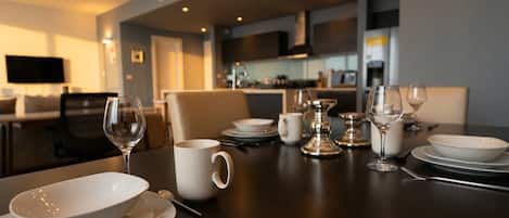 A dining table set with white dinnerware and glasses, a TV in the background, and a modern kitchen in the adjoining room.