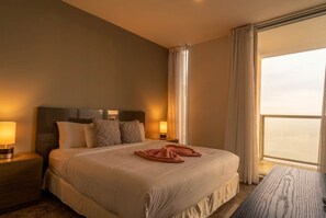 Une chambre d’hôtel avec un grand lit, deux lampes de chevet, une vue sur la mer et une fenêtre avec des rideaux.