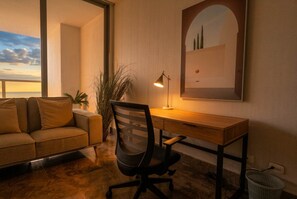 Ett rum med ett skrivbord i trä, en svart kontorsstol, en beige soffa och ett stort fönster med utsikt över himlen.