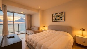 Et hotelværelse med en stor seng, et skrivebord, en stol, et natbord, en lampe, et billede og udsigt til havet.