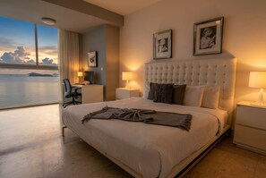 A hotel room with a large bed, two bedside tables, a desk, and a chair. There are two framed pictures on the wall, and a view of the sea through the window. - 32B Apto en Yoo con Vista al mar en Avenida Balboa (Panamá)