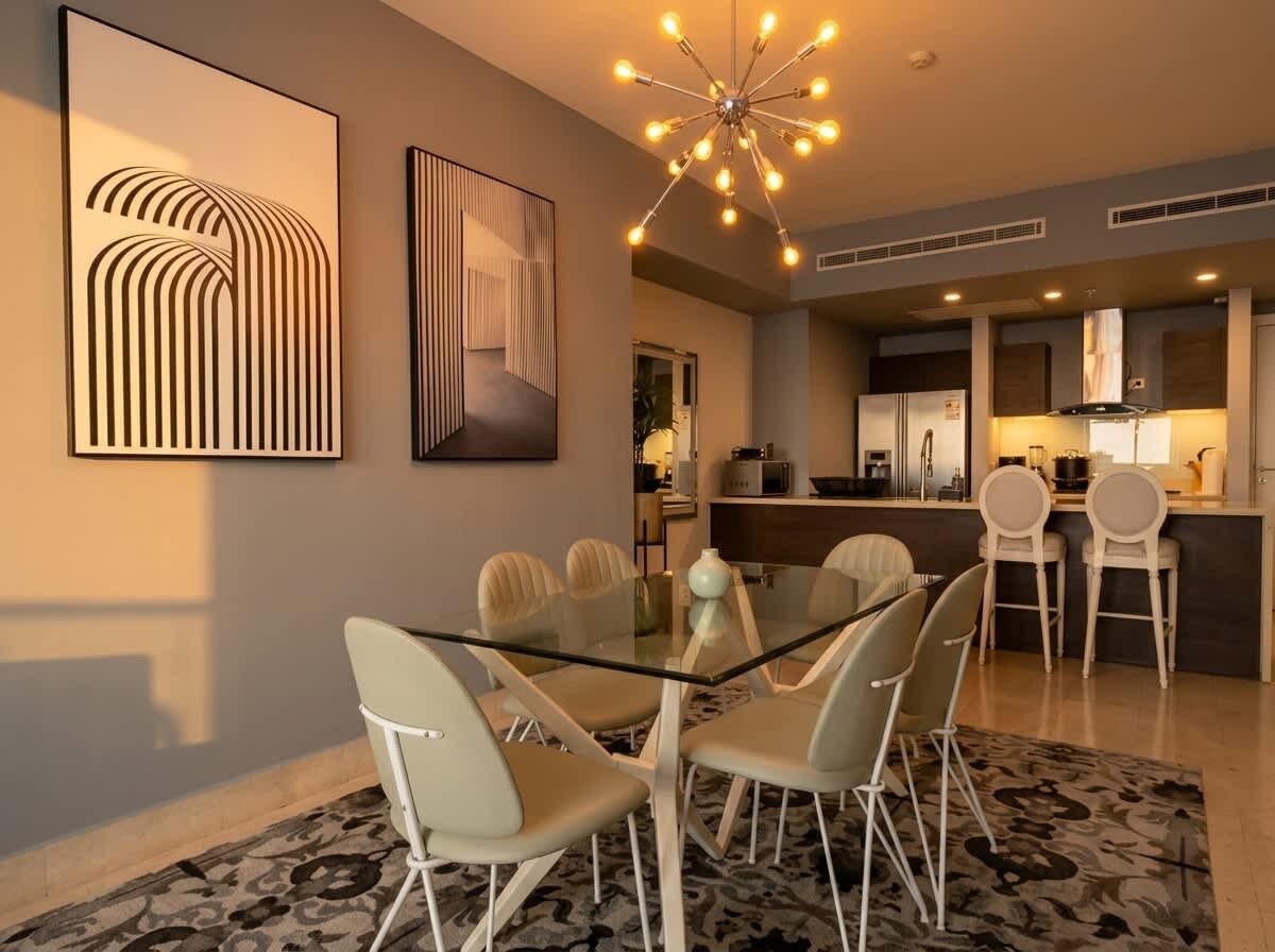 A modern dining area with a glass table and white chairs, a kitchen in the background, and a unique ceiling light fixture.