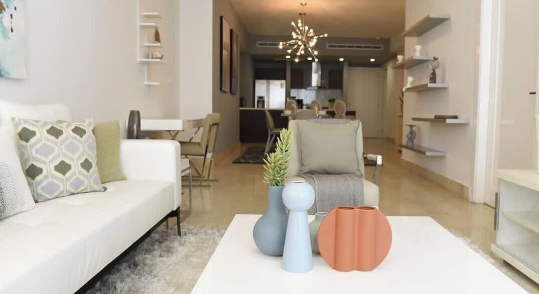 A modern living room with a white sofa, a coffee table with vases, and a dining area in the background.
