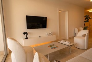 A modern living room with a flat-screen TV mounted on the wall, a white sofa, a white armchair, and a coffee table with a magazine. - Apto 61C con balcon frente al mar y 1 recamara (Panamá)