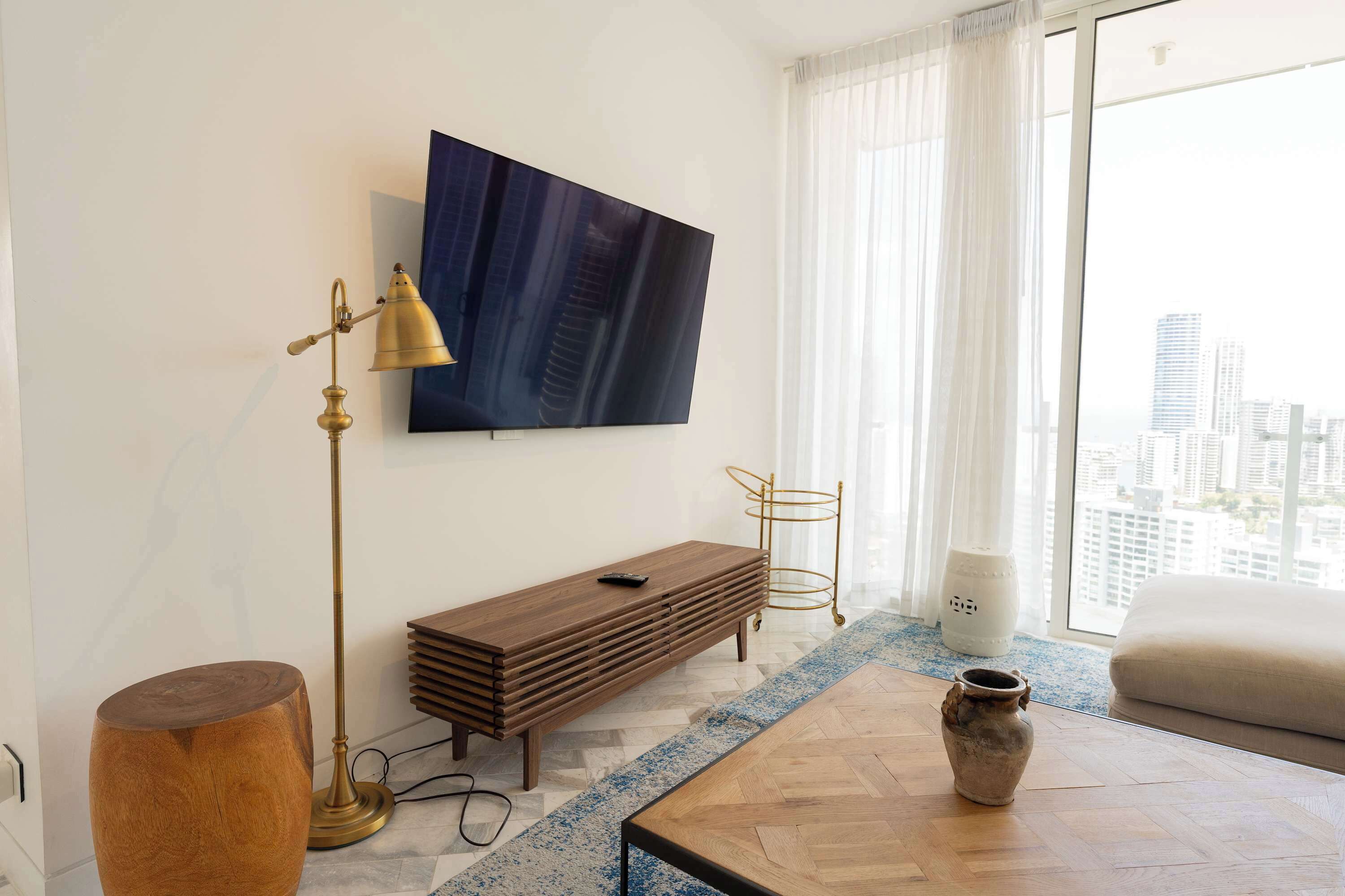 A modern living room with a TV mounted on the wall, a wooden bench, a round wooden stool, a coffee table, and a view of the city through large windows.