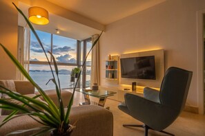 Ein modernes Wohnzimmer mit großem Fernseher, bequemen Sitzmöglichkeiten, einem Glastisch und Blick aufs Meer.