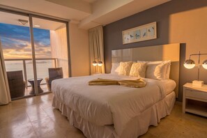 Ein Hotelzimmer mit Bett, Nachttisch, Lampe, Fenster mit Blick und einer Wand mit Kunstwerken.