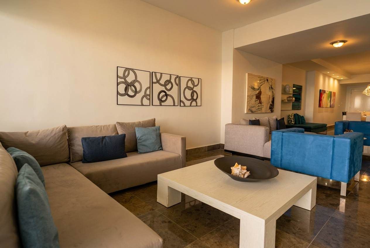 A modern living room with a beige sofa, a white coffee table, and a blue armchair. There are abstract wall art pieces above the sofa.