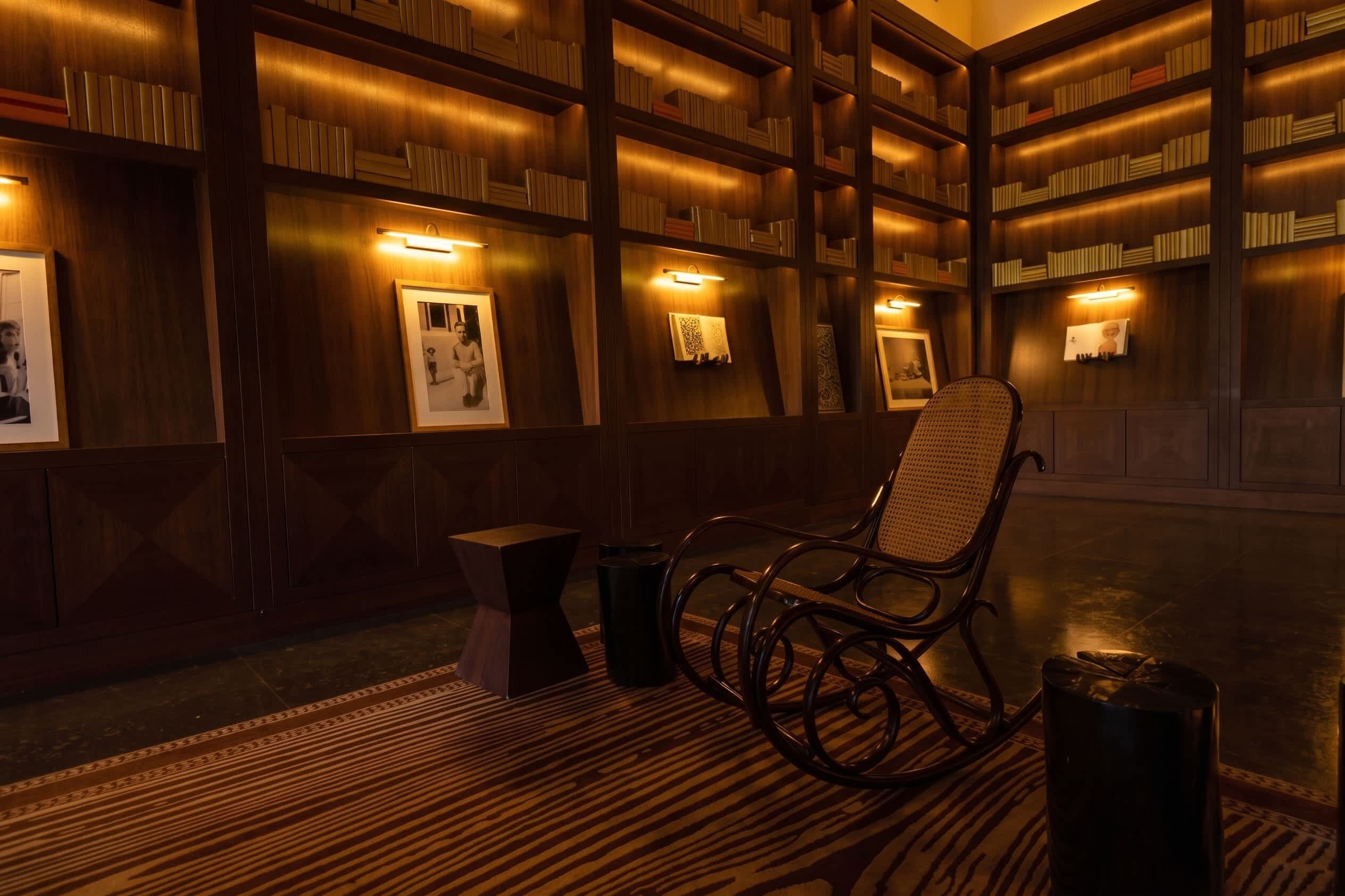 A library with bookshelves, framed pictures, a rocking chair, and a small table.