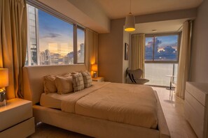 Ein Hotelzimmer mit einem großen Bett, einem Nachttisch mit Lampe, einem Sessel, einem kleinen Tisch und einem Balkon mit Blick auf die Stadt.