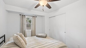 Un dormitorio con una cama, un ventilador de techo, una ventana con cortinas y una puerta.