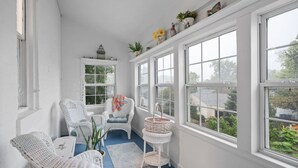 A room with white wicker furniture, a small table, a chair with a colorful blanket, and a window with a view of greenery. - Oma’s Haus - Walk to Main Street! (Saint Charles)