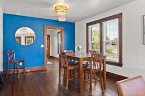A dining area with a wooden table and chairs, a blue accent wall, a round mirror, and a window with blinds. - The Barker Bungalow - Charming 3 bedroom in Historic Town near shops and dining (Saint Charles)