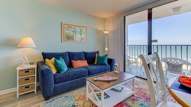 Una habitación con un sofá azul, una mesa de centro, una mesita auxiliar pequeña, una alfombra colorida y vista al mar desde un balcón.