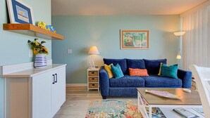 Una sala de estar con un sofá azul, una mesita, una lámpara, un tapete colorido y un cuadro en la pared.