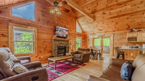 A wooden cabin interior with a fireplace, leather sofas, a television, and a dining area. - Porchside Pines։ Private Hot Tub (Pigeon Forge)