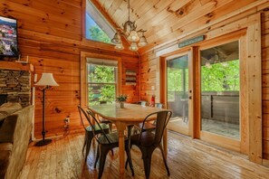 Interior de una cabaña de madera con una mesa de comedor, sillas, chimenea de piedra, televisor y ventanas amplias.