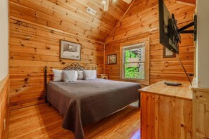Un dormitorio de cabaña de madera con una cama, un ventanal, un televisor y una mesita.