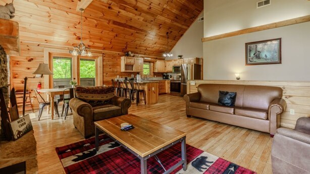 A cabin interior with a wooden ceiling, a dining area, a living room with a sofa, a kitchen, and a large window. - Porchside Pines։ Private Hot Tub (Pigeon Forge)