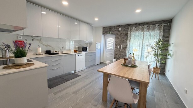 Une cuisine moderne avec des meubles blancs, une table à manger en bois et une vue sur l’extérieur par une fenêtre.