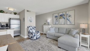 A modern living room with a grey sofa, a patterned armchair, a wooden table, and a kitchen area with white cabinets and a stainless steel refrigerator. - Charming 1 Bedroom Oceanfront Condo in Carolina Beach, perfect for 6 guests 0409 (Carolina Beach)