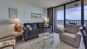 A hotel room with a grey sofa, a beige armchair, a small table, and a view of the ocean through large windows. - Experience oceanfront luxury at 0411 Seaside Sunrise, Carolina Beach! (Carolina Beach)
