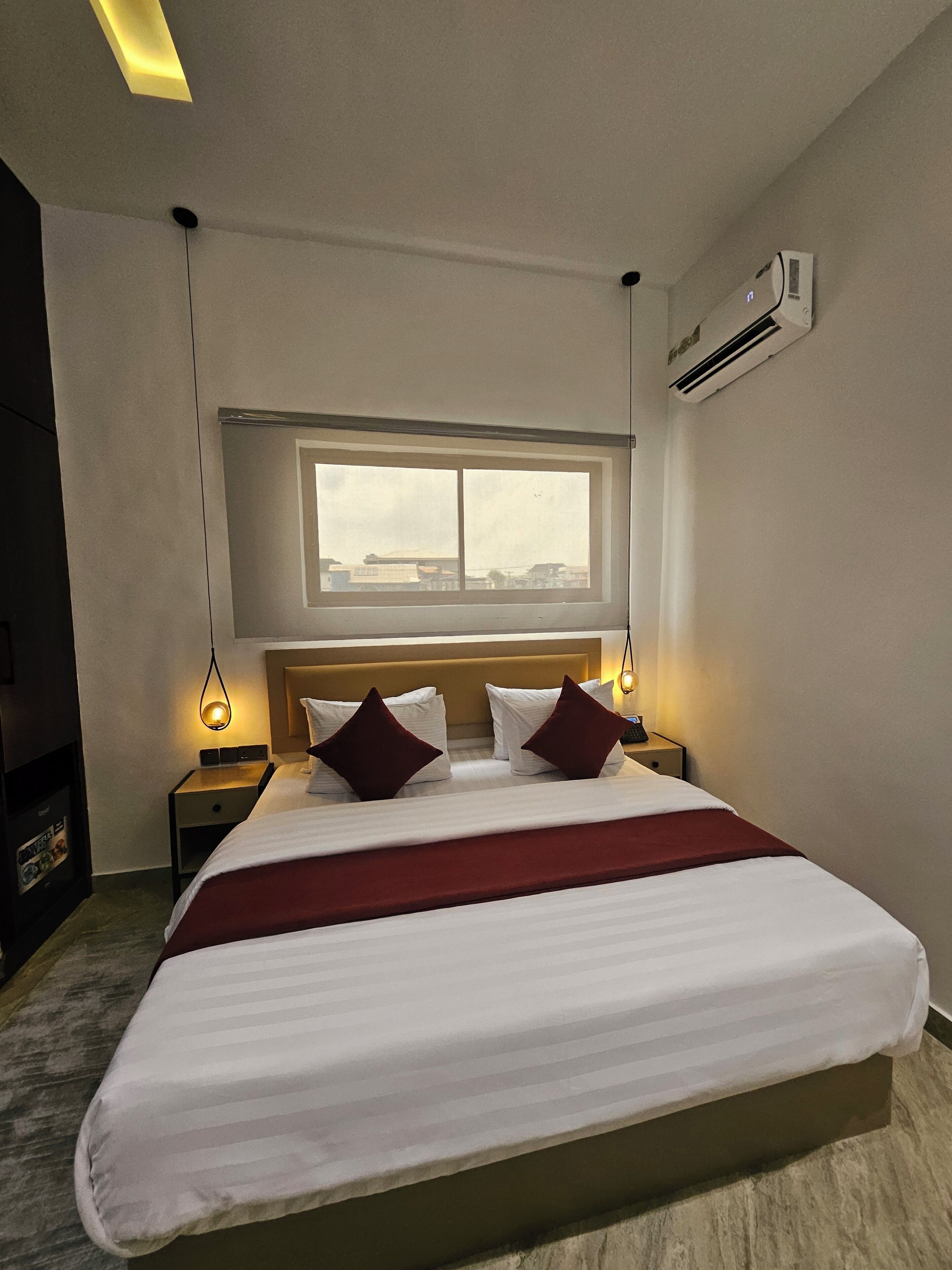 A hotel room with a large bed, a window with a view, a bedside table with a lamp, and an air conditioning unit.