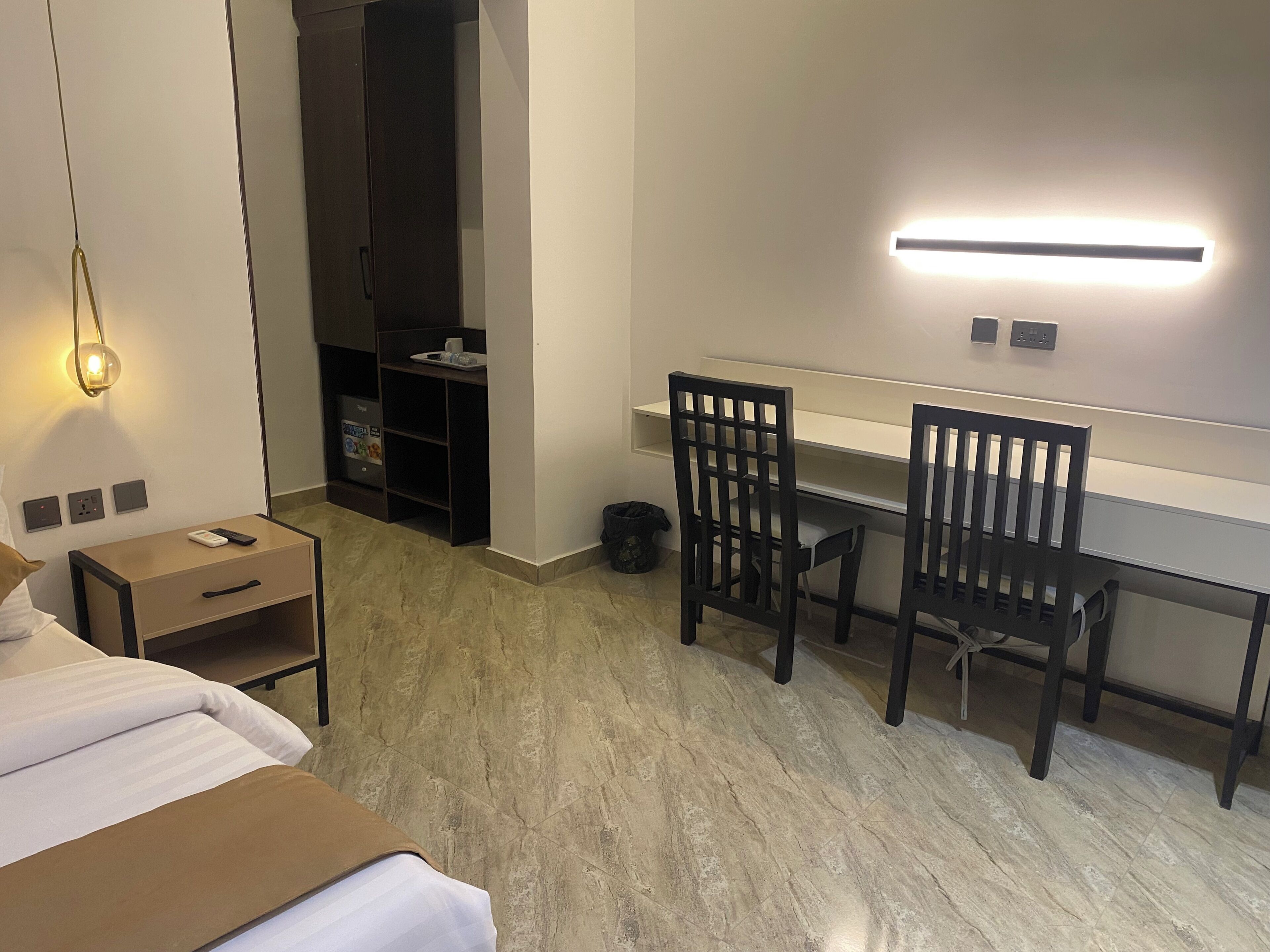 Una habitación de hotel con una cama, un escritorio con dos sillas, un armario, un cubo de basura y una lámpara fijada a la pared.