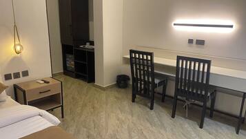 Una habitación de hotel con una cama, un escritorio con dos sillas, un armario, un cubo de basura y una lámpara fijada a la pared.