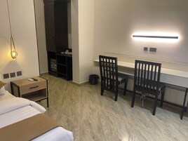 Una habitación de hotel con una cama, un escritorio con dos sillas, un armario, un cubo de basura y una lámpara fijada a la pared.