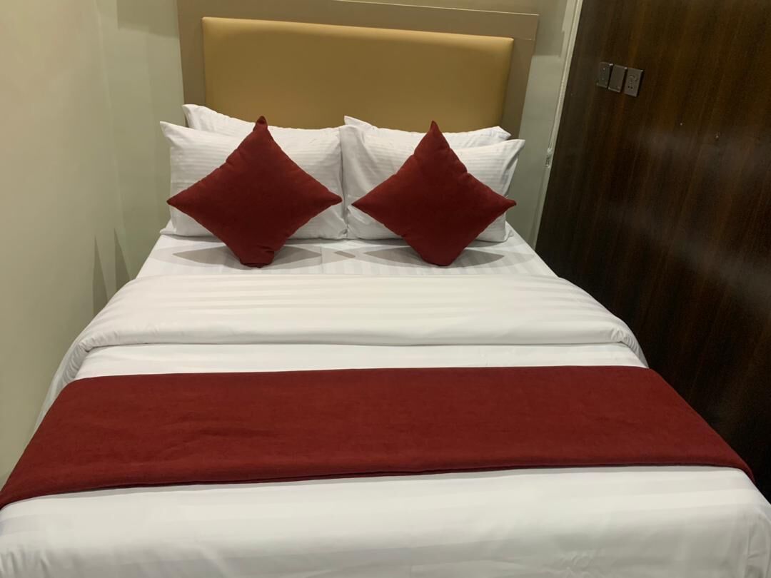 Una cama bien tendida con sábanas blancas y dos almohadas rojas, un cabecero de madera y una puerta de madera al fondo.