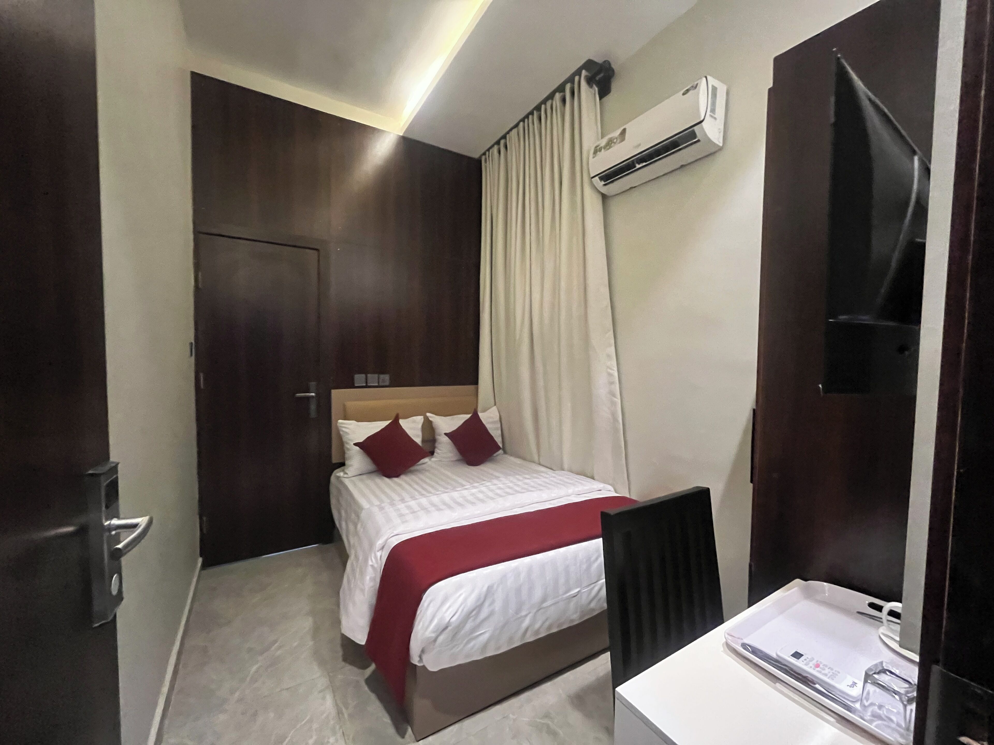 A hotel room with a bed, a desk, an air conditioning unit, a door, and a curtain.