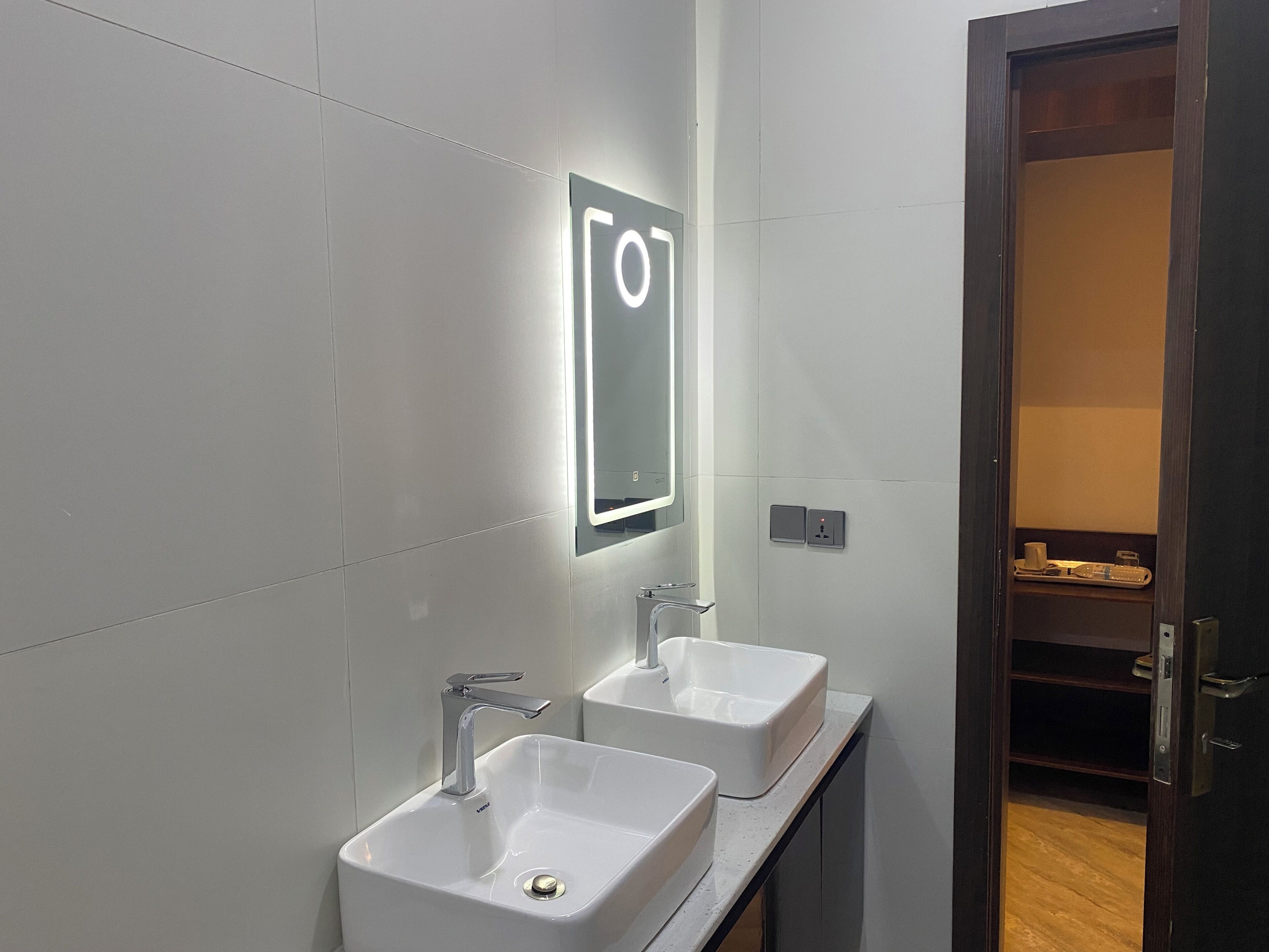 A modern bathroom with two white sinks, a mirror with illuminated signage, and a wooden door leading to a closet.