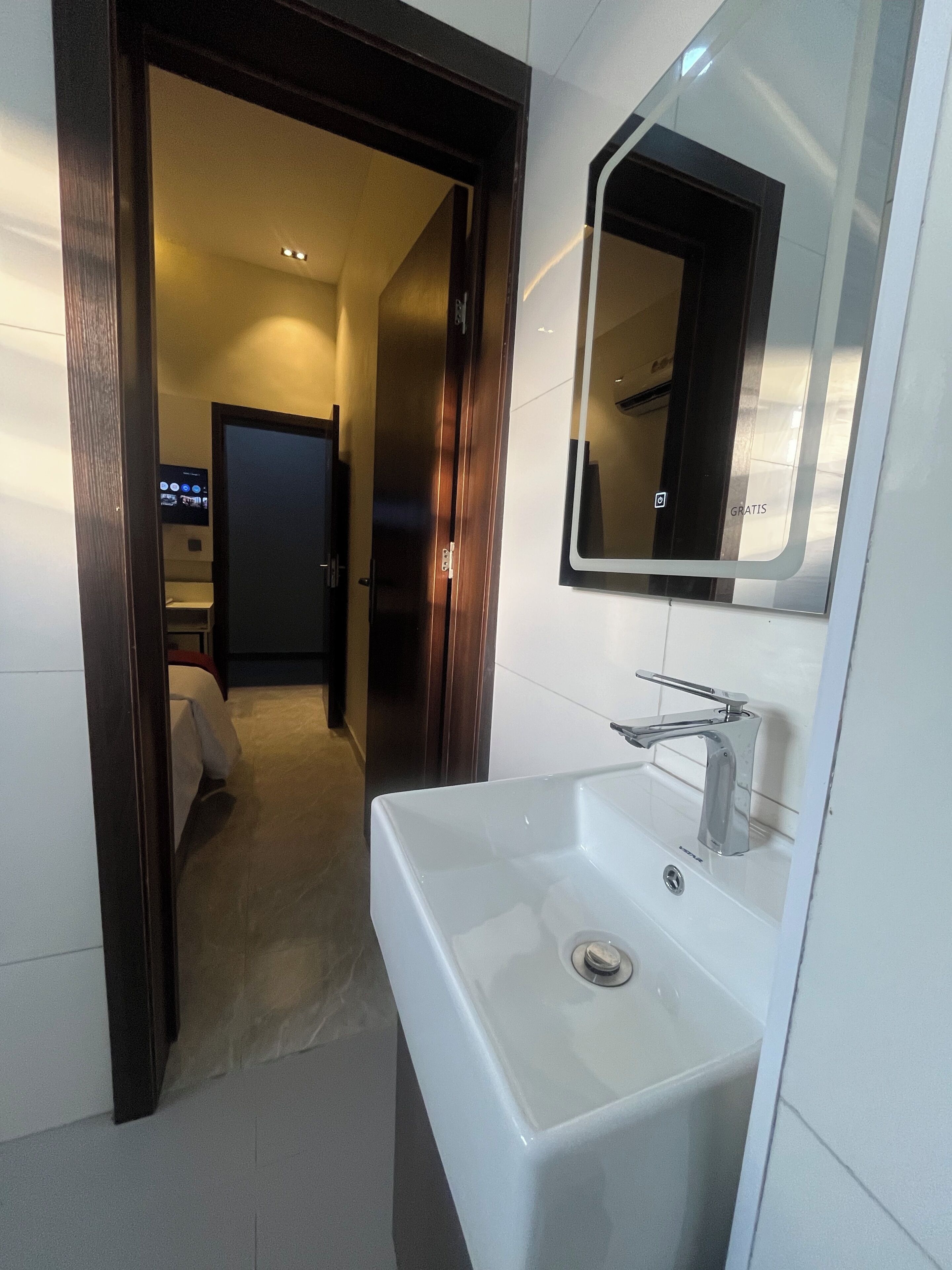 Un baño moderno con un lavamanos blanco, un espejo y una puerta que da a un pasillo.