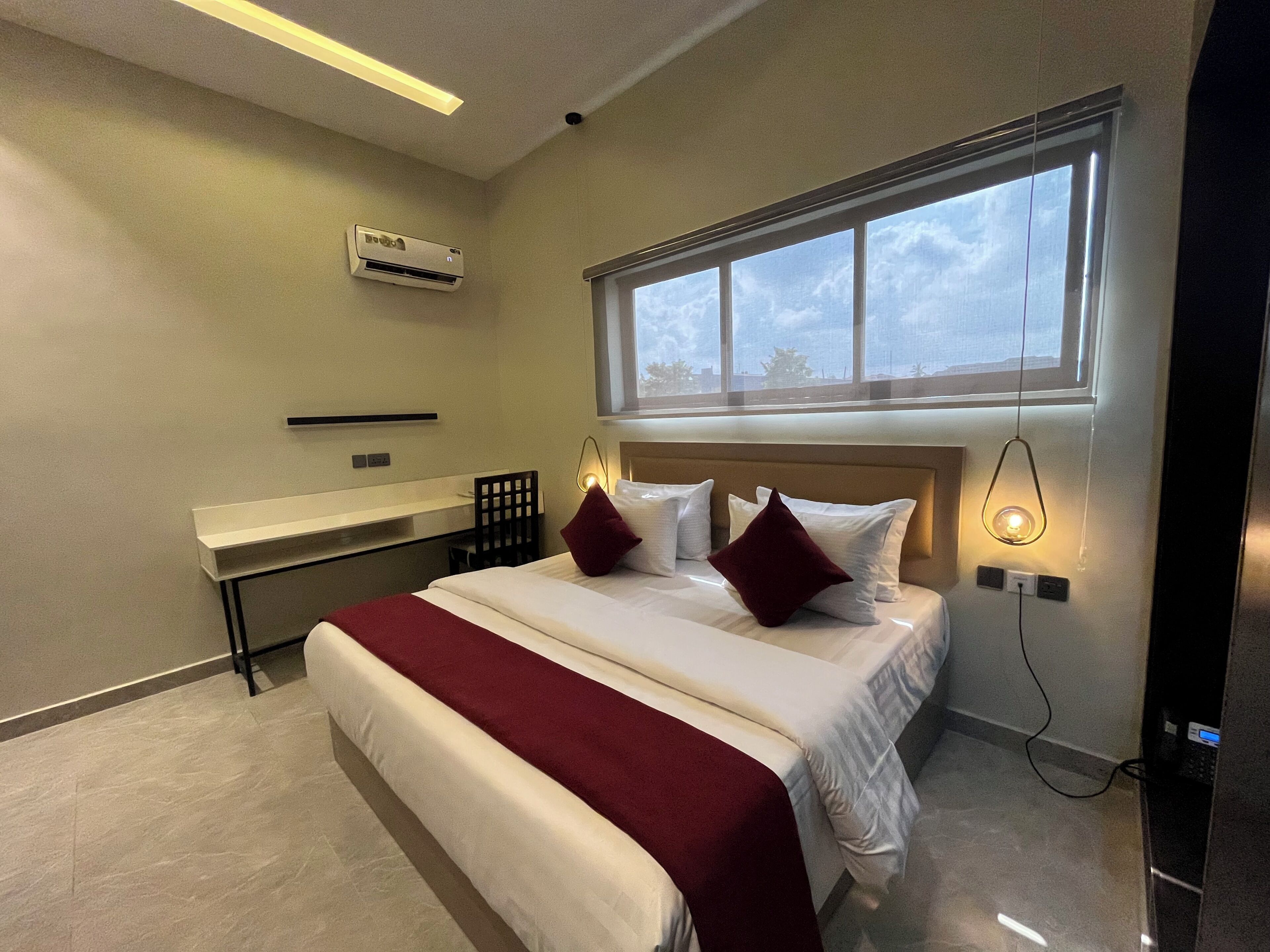 A modern hotel room with a large bed, bedside lamps, a window with a city view, and an air conditioning unit.
