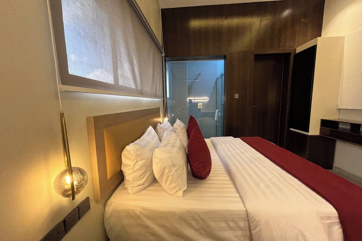 Ett hotellrum med en säng, en röd kudde, en väggmonterad lampa och ett fönster.