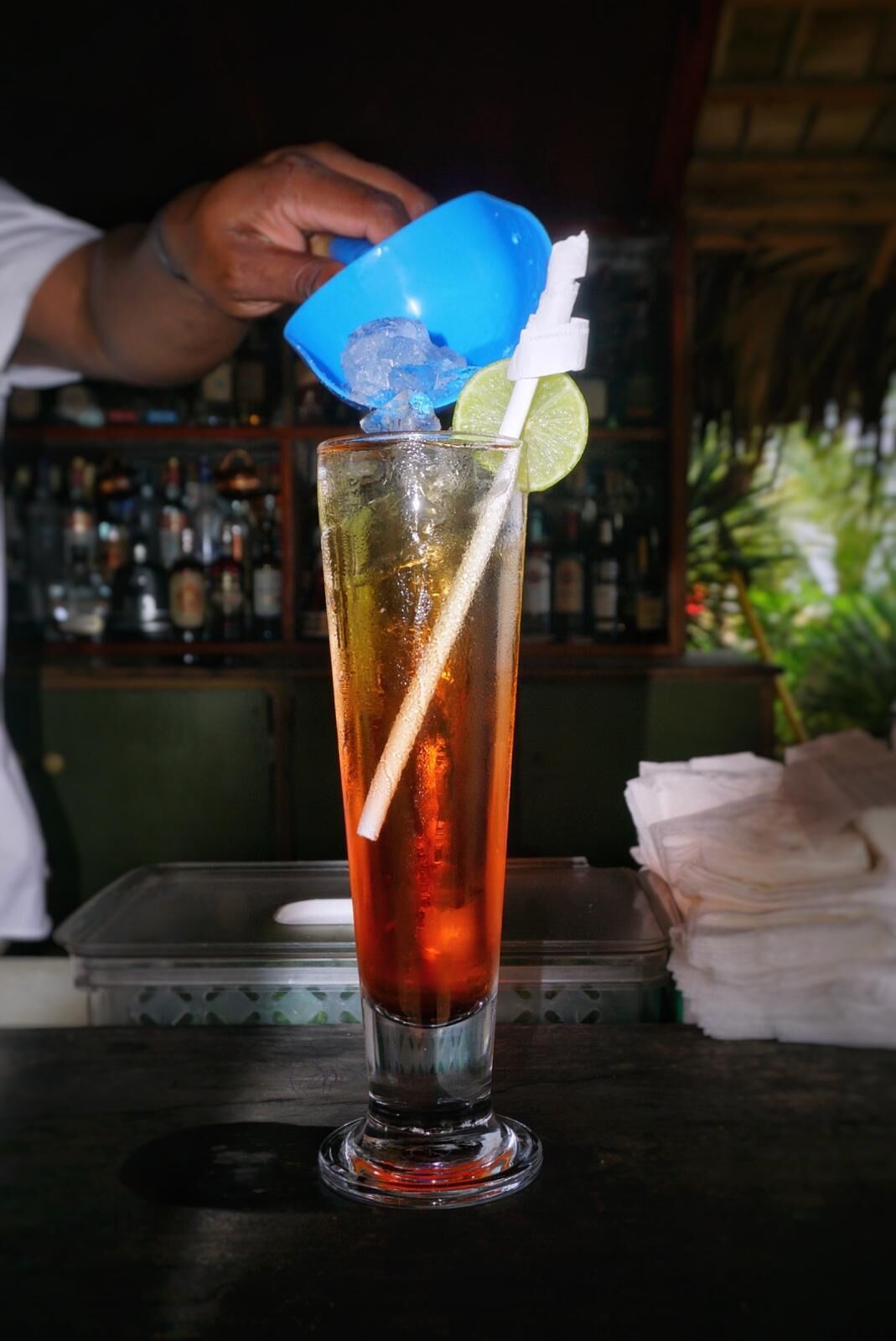 A tall glass with a straw, ice, and a lime wedge on a bar counter.