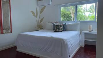 A bedroom with a bed, a nightstand, a wall-mounted air conditioner, a window with a view of greenery, and a macrame wall hanging.