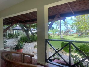 A covered outdoor area with a wooden railing, a potted plant, and a view of a garden with palm trees and a thatched-roof structure. - Hotel Boutique Casa Grande Playa Bonita (Las Terrenas)
