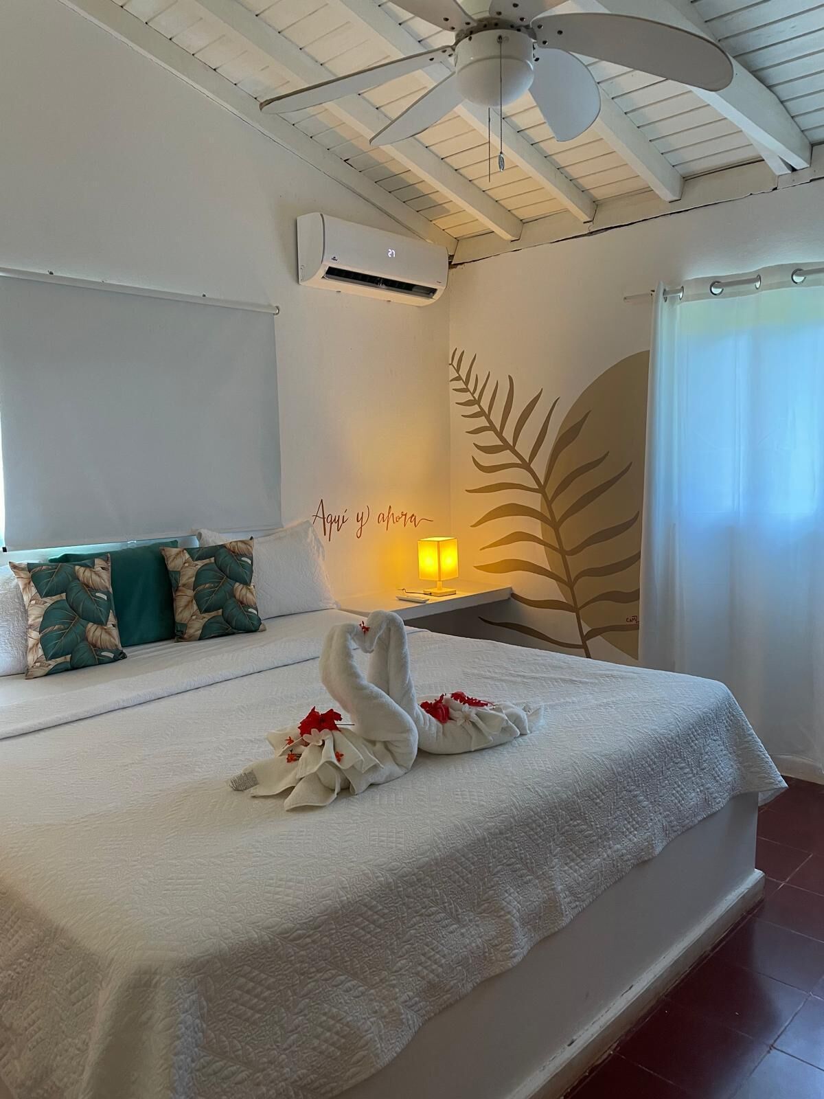 A hotel room with a bed, a ceiling fan, an air conditioning unit, a nightstand with a lamp, and a decorative wall with a leaf pattern.