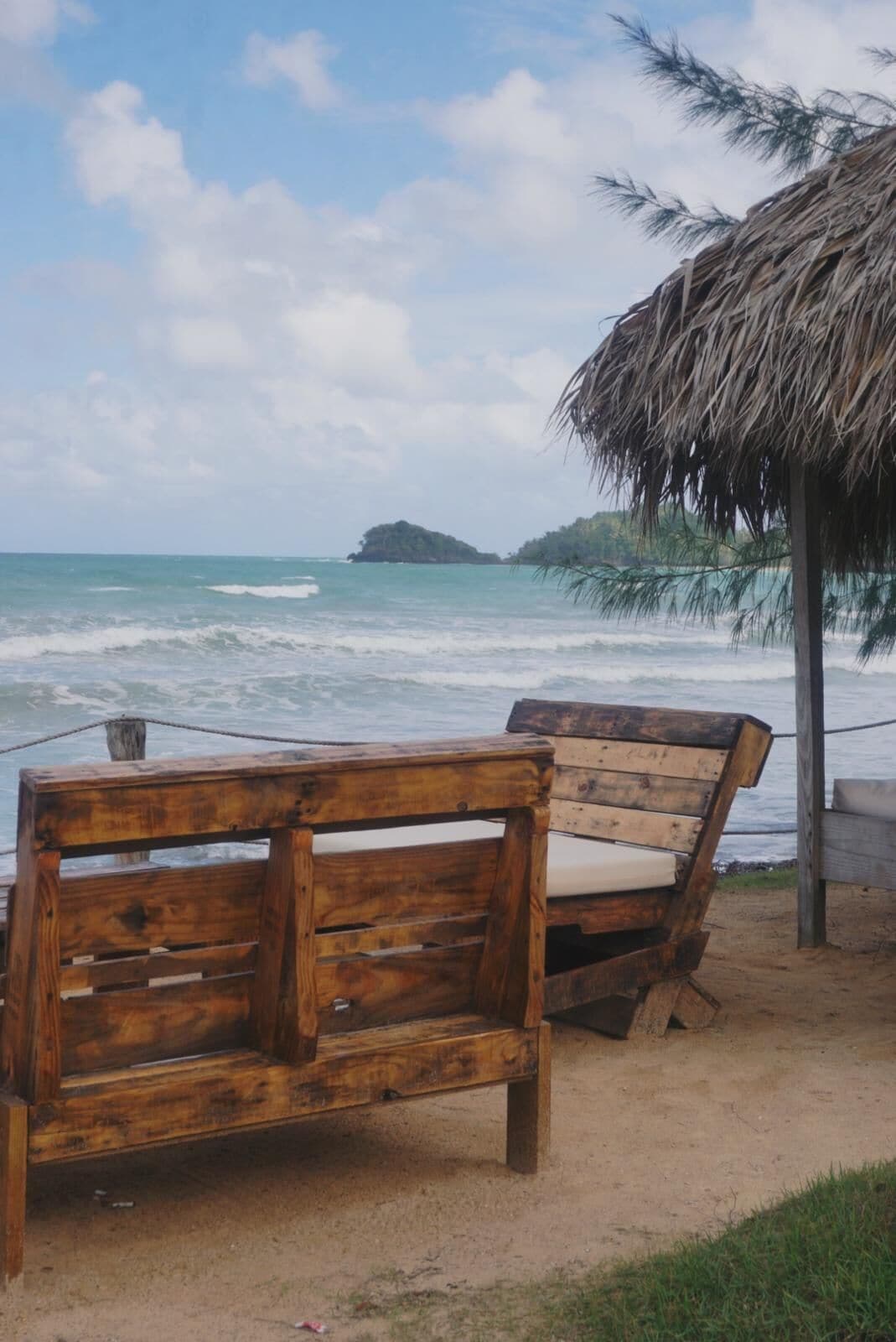 A wooden bench by the beach with a thatched roof structure nearby.