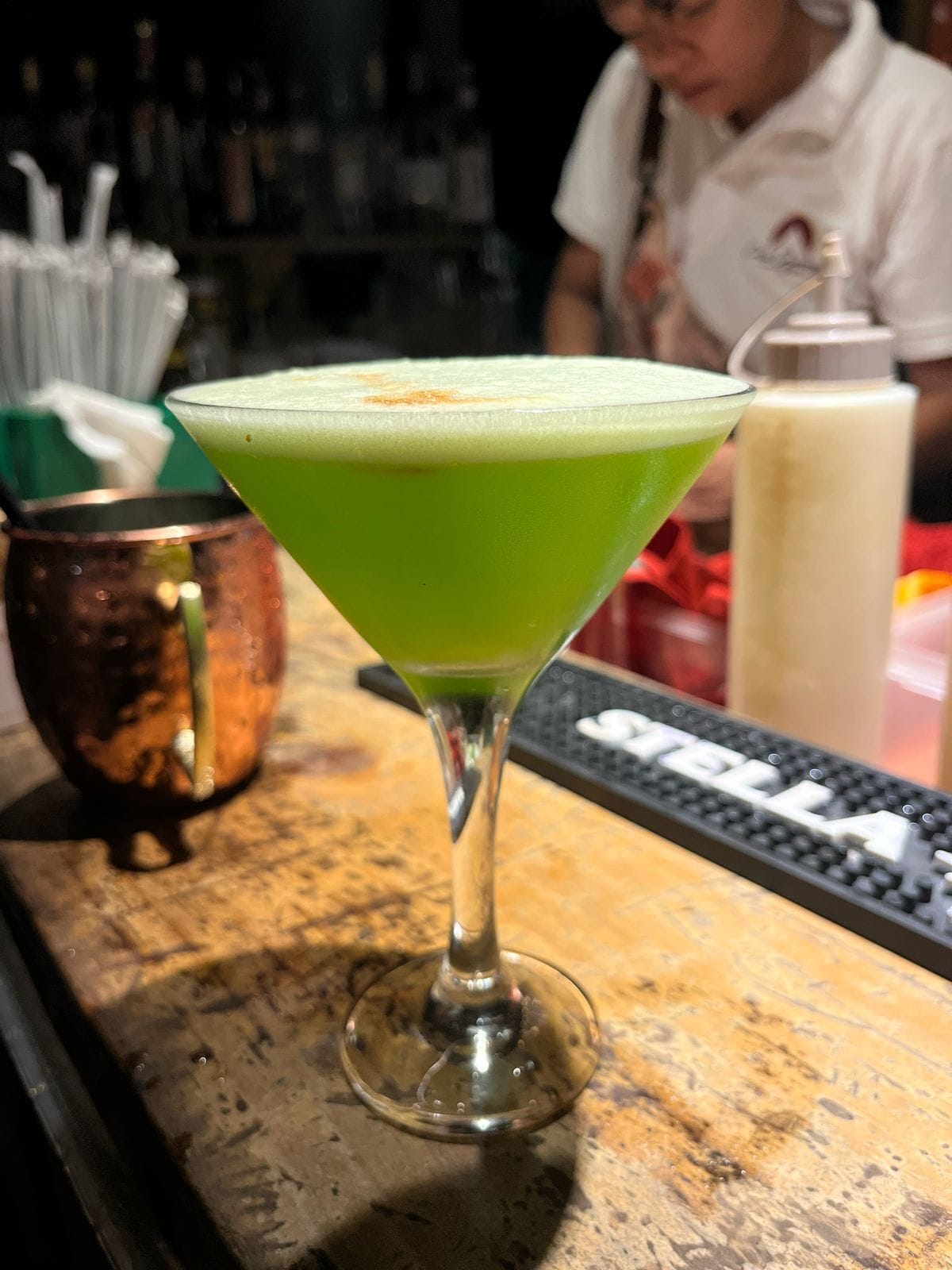 A bar setting with a green cocktail in a martini glass, a copper mug, and a keyboard in the background.